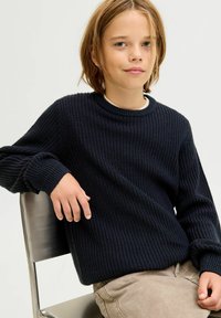 Dunkelblauer gestrickter Pullover mit strukturiertem Muster, Rundhalsausschnitt und gerippten Bündchen, getragen über einem weißen Hemd, kombiniert mit hellen Hosen.