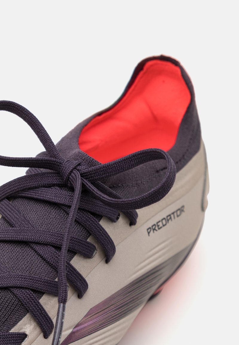 Chaussure de football grise et noire avec un tissu texturé, des lacets violets et un intérieur rouge contrastant. Présente un logo "Predator" et un design élégant.