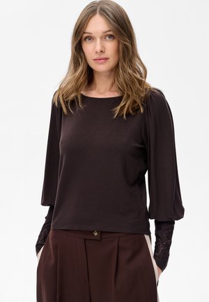 Kobieta o jasnobrązowych włosach, ubrana w ciemnobrązową bluzkę z długim rękawem i bufkami oraz ciemnobrązowe spodnie z wysokim stanem, trzyma ręce w kieszeniach.