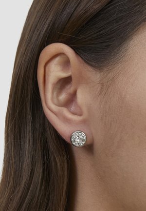Runde Ohrstecker mit einem klaren, funkelnden Stein, der von einer silberfarbenen Fassung umgeben ist, an einem Ohr eines Models zu sehen.
