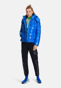 Shiny blue puffer jacket with a hood and front zipper, paired with black cargo pants, bright blue and green ankle socks, and athletic shoes.