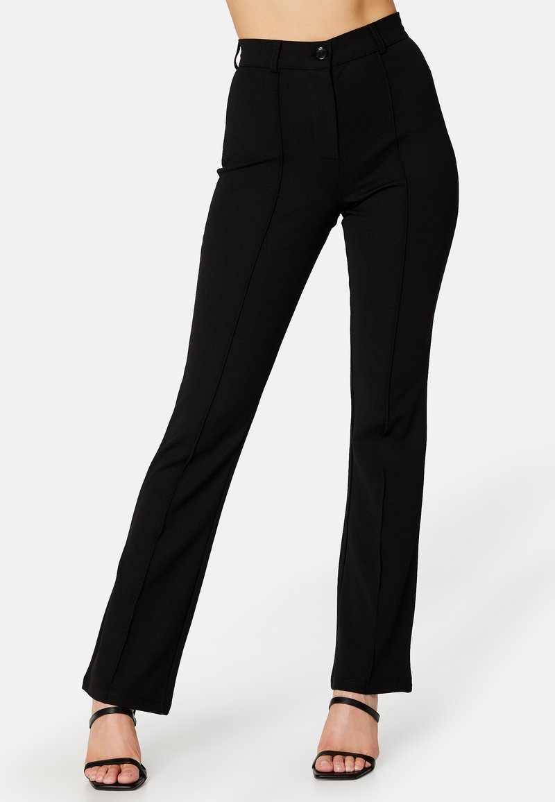 Pantalon noir taille haute, coupe slim, avec des coutures devant, porté avec des talons ouverts noirs et des brides à la cheville sur une personne debout.