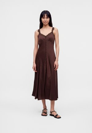 Mujer de pie que lleva un vestido midi sin mangas de color marrón oscuro con ribete de encaje y botones delanteros, combinado con sandalias planas negras.