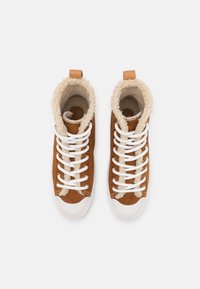 Brązowe zamszowe sneakersy typu high-top z białymi sznurówkami, podszewką z futra i kontrastującymi białymi gumowymi podeszwami. Posiadają brązową skórzaną metkę i pętelkę.