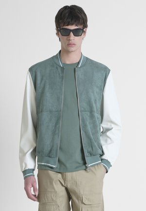 Chaqueta bomber de ante verde con mangas blancas, cierre de cremallera, puños y cuello acanalados; combinada con una camiseta verde y pantalones beiges.