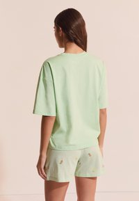 Mujer con cabello largo y oscuro, vistiendo una camiseta suelta de color verde claro y pantalones cortos a cuadros verdes con pequeños patrones de fruta, posando contra un fondo beige.