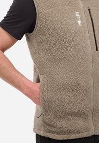 Mann trägt eine beigefarbene Fleece-Weste mit Reißverschlusstaschen über einem schwarzen kurzärmeligen Hemd, die Hand im linken Taschen.