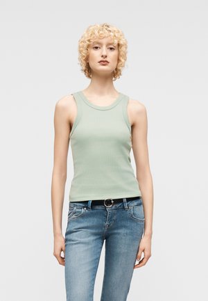 Ung person med kort, krøllet blondt hår iført en lys grøn ærmeløs top og blå jeans, stående foran en ensfarvet baggrund.