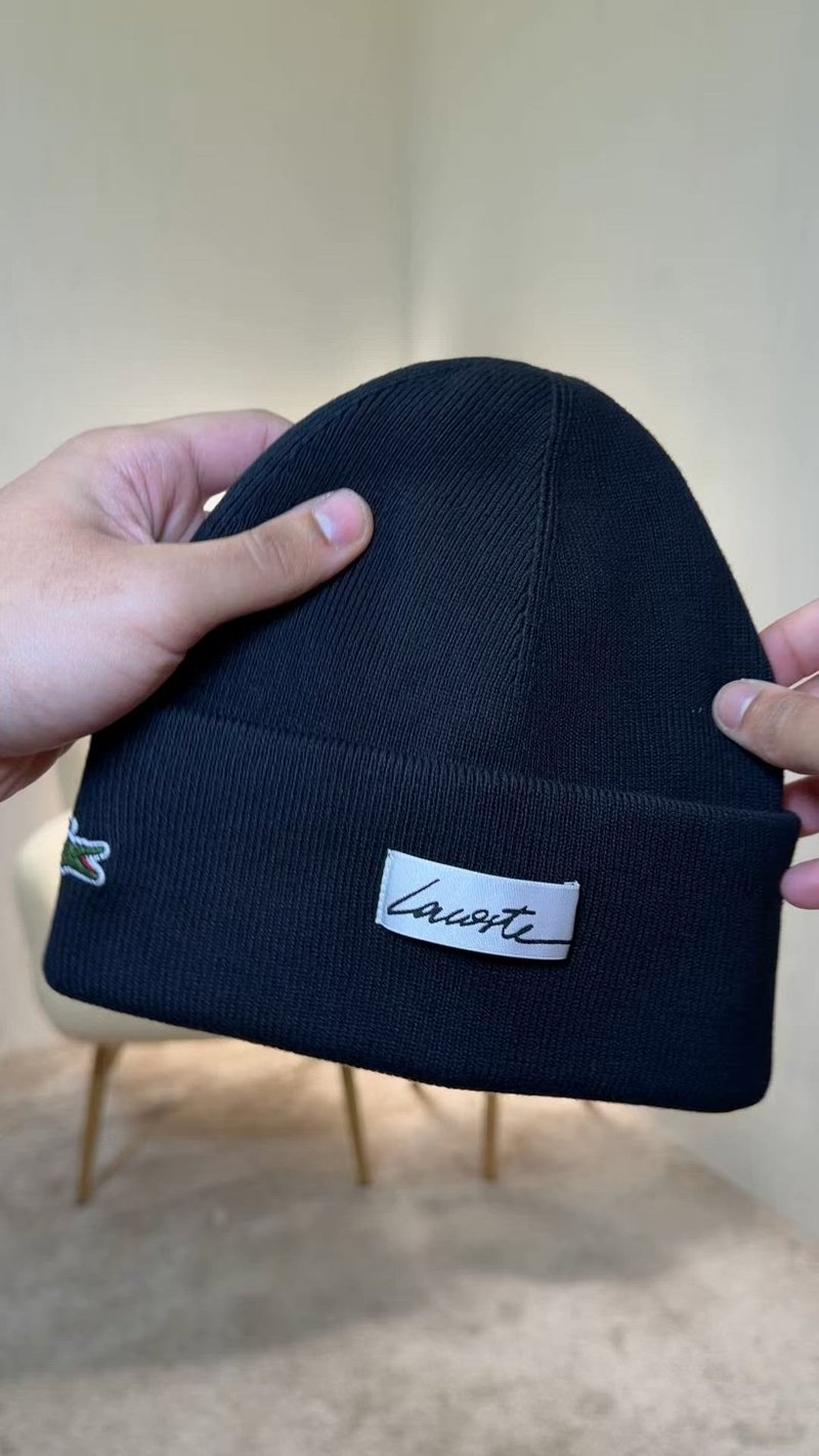 Ръце държат черна плетена шапка с бяла емблема на Lacoste и малък зелен крокодил на светъл фон.