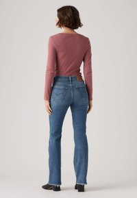 Γυναίκα με κοντά καστανά μαλλιά στέκεται γυρισμένη από την πλάτη, φοράει μακρυμάνικο πουλόβερ σε μωβ χρώμα, μπλε τζιν και μαύρα παπούτσια με τακούνι.