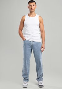 Top senza maniche bianco abbinato a jeans larghi blu chiaro e sneakers bianche con dettagli grigi, su uno sfondo grigio.
