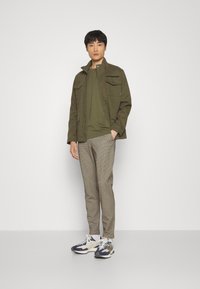 Oliwkowa kurtka z dwiema przednimi kieszeniami, w zestawie z dopasowaną zieloną koszulą i jasnobrązowymi spodniami w kratę. Białe i granatowe sneakersy.