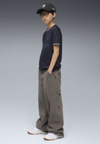 T-shirt i marineblå med hvid kant, cargo grå bukser og hvide sneakers. Accessories inkluderer en sort kasket og runde briller.