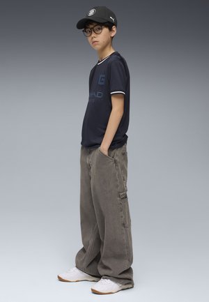 T-Shirt in Marineblau mit weißen Akzenten, Cargo-graue Hose und weiße Sneakers. Die Accessoires umfassen eine schwarze Mütze und runde Brillen.