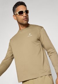 Long-sleeve, light brown t-shirt with a round neckline and a white logo. Styled with light-colored sunglasses against a gray background.