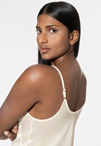 Crème-kleurige satijnen cami met verstelbare bandjes en gouden hardware, die een gladde textuur en een aangesloten ontwerp laat zien.