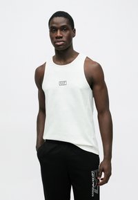 Mladý muž v bielom tielku EA7 Emporio Armani bez rukávov a čiernych nohaviciach so zodpovedajúcim logom stojí pred jednoliatym svetlým pozadím.