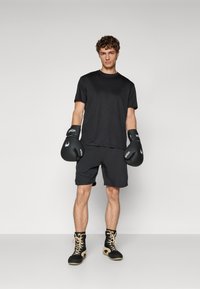 T-shirt de sport noir et shorts noirs avec des gants de boxe noirs. Le modèle porte des chaussures de sport montantes noir et beige. Tissu lisse et respirant.