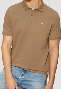 Polo shirt marron à manches courtes avec un tissu texturé, col à deux boutons et petit logo sur la poitrine gauche, assorti à un jean en denim bleu clair.