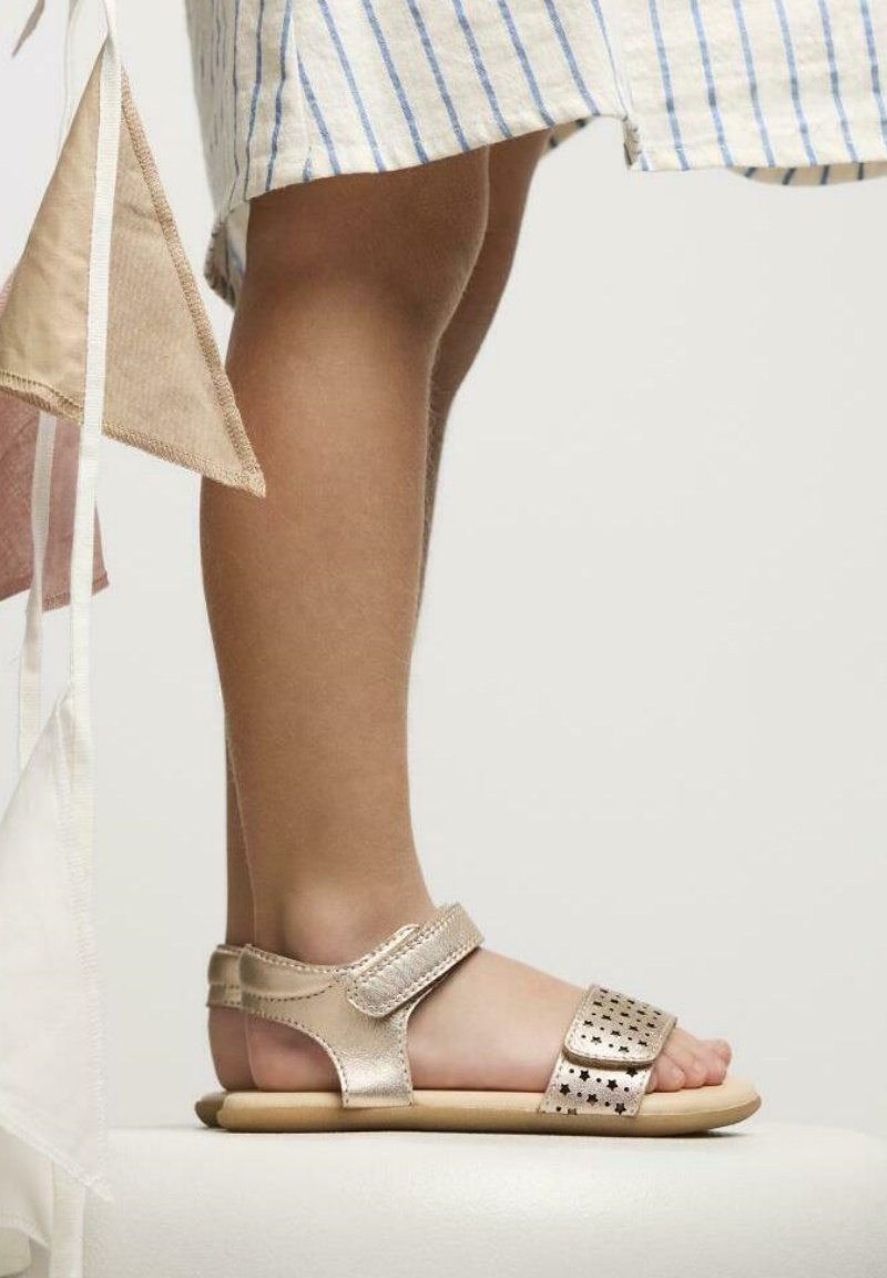 Sandal in oro rosa metallizzato con cinturino regolabile, design a stella perforata e suola piatta beige. Indossata dal piede di un bambino dalla pelle chiara.