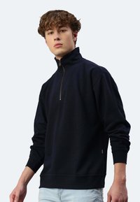 Schwarzer Fleece-Pullover mit hohem Kragen, halbem Reißverschluss, gerippten Bündchen und einfarbiger Textur. Besitzt ein kleines Logo an der Seite.