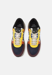 Scarpe sportive multicolori con tomaia in rete nei colori blu navy, bordeaux, arancione e giallo. Presentano lacci spessi e una suola testurizzata. Logo del marchio visibile.