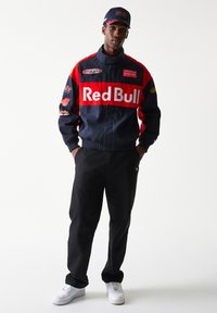 Κόκκινο και ναυτικό μπλε σακάκι Red Bull Racing με λογότυπα, μαύρο παντελόνι και λευκά αθλητικά παπούτσια. Το σακάκι διαθέτει φερμουάρ και ψηλό γιακά.