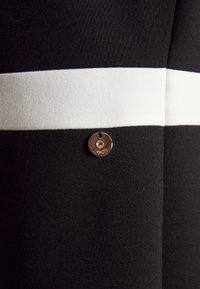 Tissu noir avec une bande blanche horizontale et une petite étiquette ronde en métal doré attachée, gravée du mot "LIU•JO".