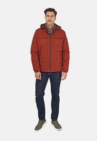 Chaqueta acolchada roja con capucha, que cuenta con dos bolsillos en el pecho, cierre de cremallera y ribete gris en contraste. Llevada con jeans oscuros y zapatillas verdes.