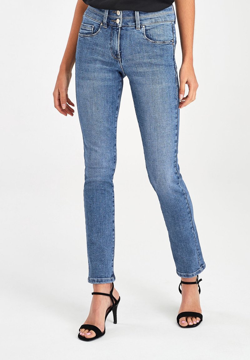 Vaqueros ajustados de denim azul con diseño de cinco bolsillos, cierre de botón y ligeras marcas de uso; combinados con sandalias de tacón alto negras con tiras.