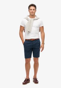 Navyblauwe katoenen shorts, gecombineerd met een witte T-shirt met ronde hals. Een lichtgrijze trui hangt over de schouders. Bruine leren riem en donkere schoenen.