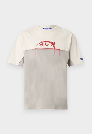 Mężczyzna z krótkimi włosami, ubrany w beżowo-szarą koszulkę z nadrukiem, różowe szorty, czarne skarpetki i czarne trampki, stoi z rękami w kieszeniach.