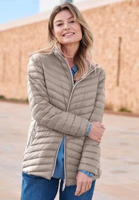 Lachende vrouw buiten in een lichtgrijze gewatteerde jas over een blauwe spijkerblouse en jeans, met haar armen gekruist voor zich.