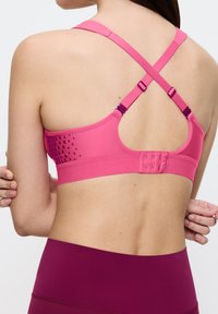 Różowy sportowy stanik z krzyżującymi się ramiączkami, gładką teksturą, regulowanymi zapięciami oraz wzorzystym bocznym wzorem przechodzącym w jednolity tył.