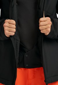Zwarte jas met een volledige rits en drukknopen, die een textuurlaag aan de binnenkant laat zien met een oranje buitenlaag die aan de onderkant zichtbaar is.
