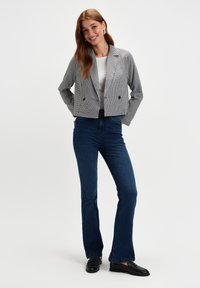 Młoda kobieta uśmiechająca się, ubrana w w kratkę marynarkę, białą bluzkę, ciemnoniebieskie dżinsy i czarne loafersy, stojąca z rękami w kieszeniach na białym tle.