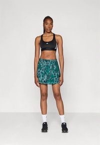 Ženska stoji oblečena v črno športno srajco, zelene kratke športne hlače s potiskom, bele nogavice do gležnjev in črne tekaške čevlje na enobarvni podlagi.