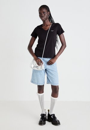 Femme avec des cheveux tressés portant un t-shirt noir à col en V, un short en denim long bleu clair, de hautes chaussettes blanches en tricot, des chaussures à plateforme noires et un petit sac en bandoulière en argent.