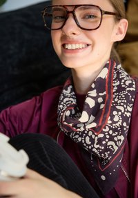 Écharpe en soie tissée de couleur violet foncé avec des motifs floraux blancs et une bordure rouge ; portée avec une veste bordeaux, de coupe décontractée.