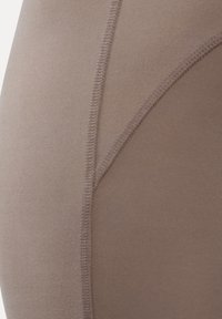 Close-up van taupe stof met zichtbare gebogen stiksels, die de textuur en details van het materiaal tonen.