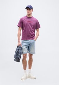 Νεαρός άνδρας που φοράει μωβ T-shirt Calvin Klein, ανοιχτόχρωμα τζιν σορτς, λευκά αθλητικά παπούτσια, μπλε σκούφο, κρατώντας ένα μπλε πουκάμισο με καρό μοτίβο σε ένα χέρι.