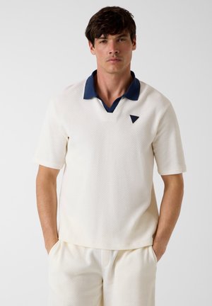 Man draagt textuur witte korte mouw shirt met marineblauwe kraag en bijpassende witte broek, handen in de zakken, neutrale achtergrond.