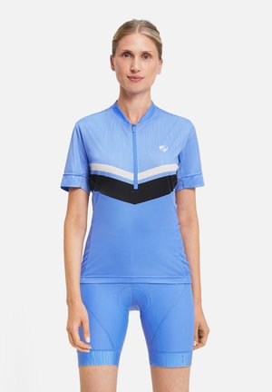 Woman wearing light blue cycling jersey with black and white chevron stripes and matching padded cycling shorts, standing against white background.