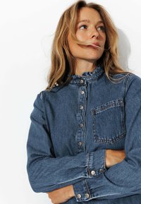 Chemise en denim bleu foncé avec un col froncé, boutonnière à l'avant et une poche poitrine unique. Présente des boutons-pression argentés et des manches longues.