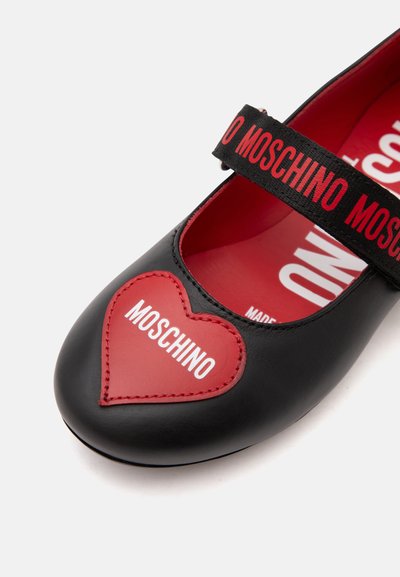 Chaussure Mary Jane noire avec patch cœur rouge portant la mention « Moschino » sur l’orteil et bride noire avec texte rouge « Moschino » sur le dessus.