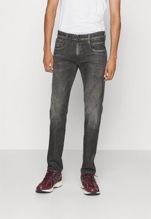 Mężczyzna w wyblakłych czarnych jeansach, białej koszulce i bordo sportowych butach, stojący na tle jednolitego jasnego tła.