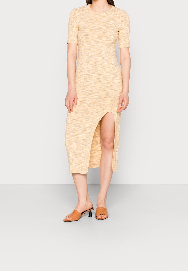 Femme portant une robe midi beige à manches courtes avec une fente haute devant à la cuisse et des sandales à talons beiges, debout sur un fond uni.