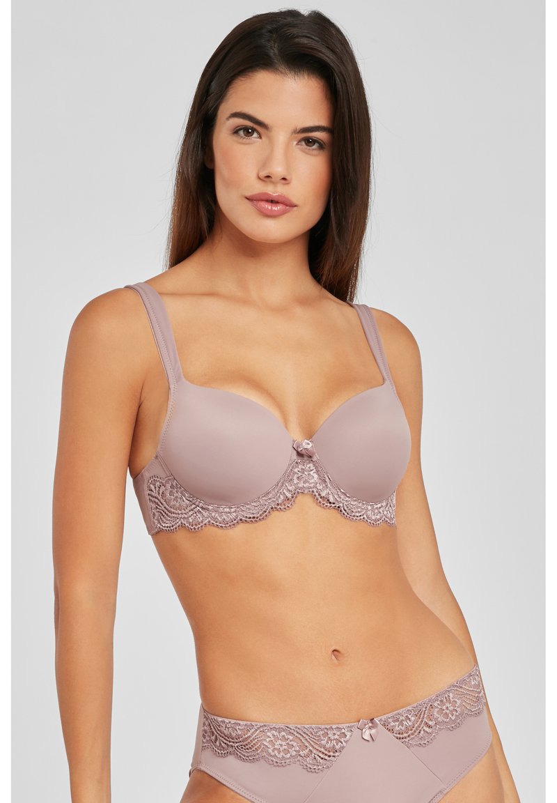 Licht mauve beha met kantafwerking en een decoratieve strik in het midden. De cups zijn glad, en de bandjes zijn breed voor ondersteuning. Bijpassende kanten slipjes zijn zichtbaar.