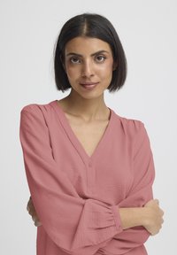 Rosafarvet bluse med V-hals, knappedetalje og vide ærmer. Tekstureret stof med diskret plissering ved manchetterne.