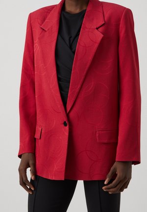 Czerwona marynarka z subtelnymi okrągłymi wzorami, pojedynczy czarny guzik, noszona na czarnej bluzce, w połączeniu z czarnymi spodniami, pokazana od ramion do bioder.